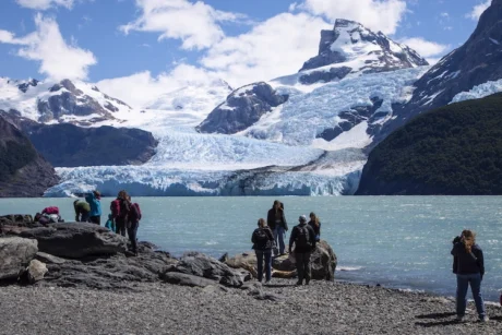 Un grupo de personas, vestidas con ropa de abrigo, se encuentran en una orilla rocosa y de grava, con la vista puesta en un gran glaciar que desciende por una montaña nevada. El glaciar, de un color azul intenso, termina en un lago de aguas color turquesa. El cielo es parcialmente nublado, y se pueden ver más picos nevados a los lados. La escena transmite una sensación de asombro y exploración de un paisaje natural majestuoso y frío.