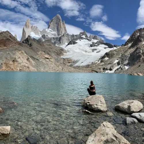 Aguas turquesas de la Laguna de los Tres con el Monte Fitz Roy de fondo y una excursionista sentada en la orilla, El Chaltén.